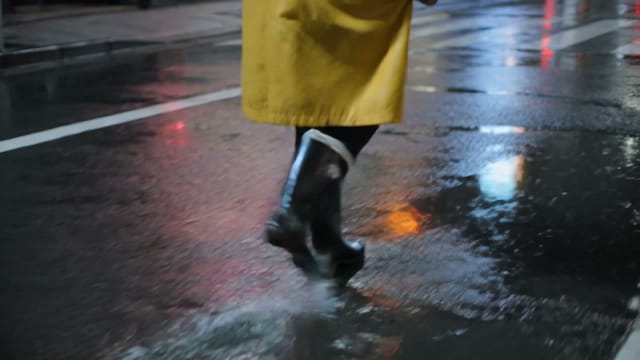 Handheld shoulder-cam drift following a woman in a yellow raincoat crossing a rain-soaked Tokyo street at night, neon reflections in puddles, umbrellas passing in the foreground, soft focus falloff.