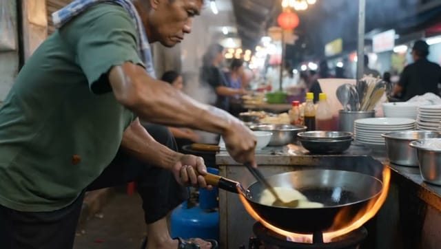 A street food vendor in Bangkok flips noodles in a flaming wok at night, fire leaping up as he tosses them high. Handheld close-up, steam and smoke catching the orange glow of string lights, busy crowd blurred behind. Documentary realism, warm tones. Audio: sizzling oil, clanging metal spatula, chatter and scooter horns in the background.