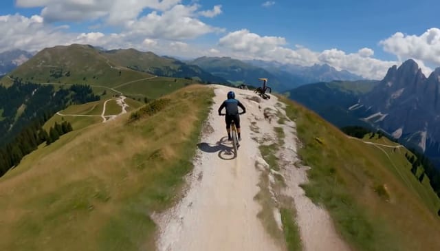 A mountain biker bombs down a narrow ridge trail in the Dolomites, launching off a rock drop and landing in a spray of dust. Low FPV drone chases right behind the rear wheel, then pulls up and away to reveal the valley. Midday sun, crisp contrast, high-energy pacing. Audio: tires crunching gravel, wind roar, the rider's sharp exhale on landing.