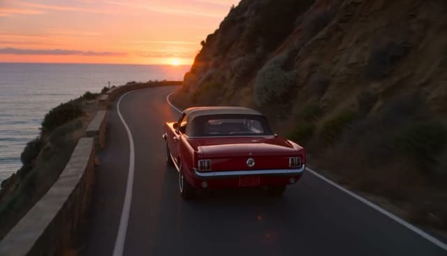 A red vintage convertible drives along a winding coastal road at sunset, cliffs and ocean below, drone shot slowly following from behind. Audio: engine hum, wind, waves far below.