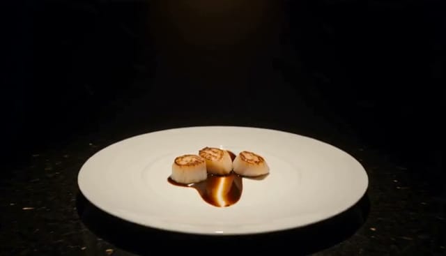 0-5s: A chef sets an empty plate on a counter and picks up a spoon. 5-15s: Slow push-in as sauce is drizzled across the plate in one line. 15-25s: A hand places three scallops down, one at a time. 25-30s: Pull back to a wide shot of the finished plate under a warm overhead light. <Kitchen room tone, drizzle stretched long>